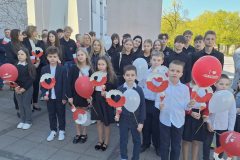 Grupa dzieci, młodzieży i dorosłych stoi na zewnątrz przed budynkiem podczas uroczystości o charakterze patriotycznym. Uczestnicy są ubrani odświętnie – głównie w ciemne stroje z białymi elementami – i trzymają biało-czerwone dekoracje oraz czerwone i białe balony. Wiele osób patrzy w stronę aparatu. W tle widać budynek oraz zielone drzewa oświetlone słońcem.