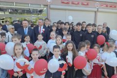 Grupa dzieci, młodzieży i dorosłych stoi na zewnątrz przed budynkiem podczas uroczystości o charakterze patriotycznym. Uczestnicy są ubrani odświętnie – głównie w ciemne stroje z białymi elementami – i trzymają biało-czerwone dekoracje oraz czerwone i białe balony. Wiele osób patrzy w stronę aparatu. W tle widać budynek oraz zielone drzewa oświetlone słońcem.