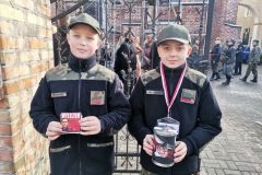 Dwaj harcerze stoją przed tłumem składającym kwiaty pod tablicą pamięci.