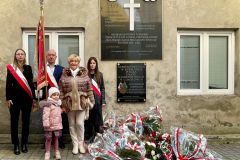 Grupa osób ze sztandarem przed tablicą pamięci; na ziemi wieńce w barwach Polski.