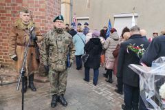 Dwaj mężczyźni w mundurach stoją przed tłumem składającym kwiaty pod tablicą pamięci.