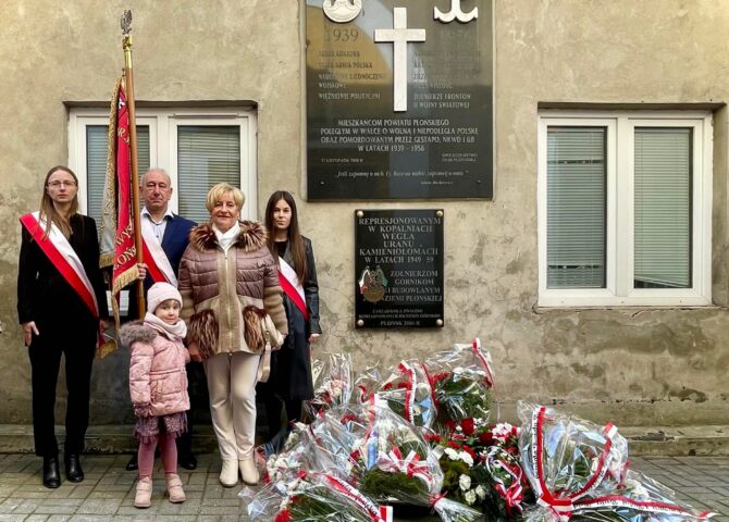 Grupa osób z flagą przed tablicą pamięci; na ziemi wieńce w barwach Polski.